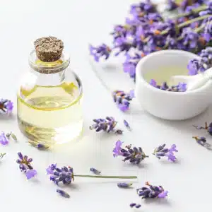 A clear glass bottle with cork contains yellow liquid beside scattered purple lavender flowers. A white ceramic bowl filled with similar liquid and accompanied by a white spoon complements the OsiMagnesium – Good Night Melatonin Body Lotion (100ml).
